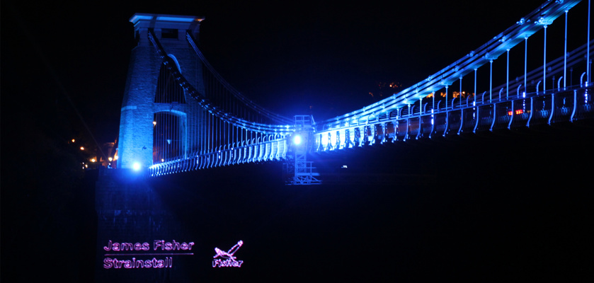 The Clifton Suspension Bridge in Bristol dramatically illuminated in blue light – with a prominent James Fisher Strainstall logo.