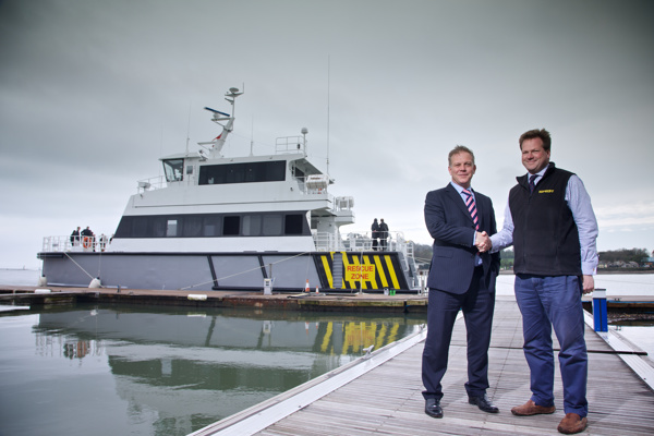 Representatives from James Fisher and Supacat shaking hands to celebrate the purchase of a multi-purpose vessel (SMV 24) which is in the background