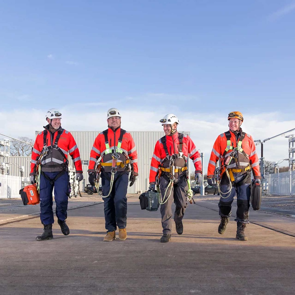 Four high voltage engineers walking in a line towards the camera with HV equipment in the background
