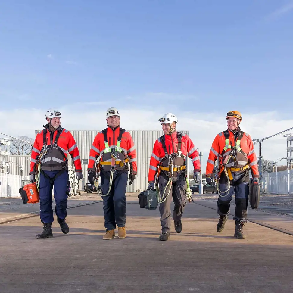 Four high voltage engineers walking in a line towards the camera with HV equipment in the background